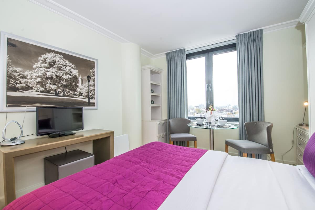 A hotel room with a bed, a desk with a computer, a television, two chairs, a small table with a cup and saucer, and a window with curtains.