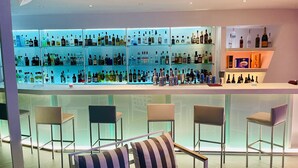 A modern bar area with multiple shelves of liquor bottles, bar stools, and a seating area with striped cushions. - Loucas On The Cliff (Santorini)