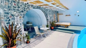 A sunlit patio with a pool, a seating area with chairs and a table, and a bed. - Loucas On The Cliff (Santorini)