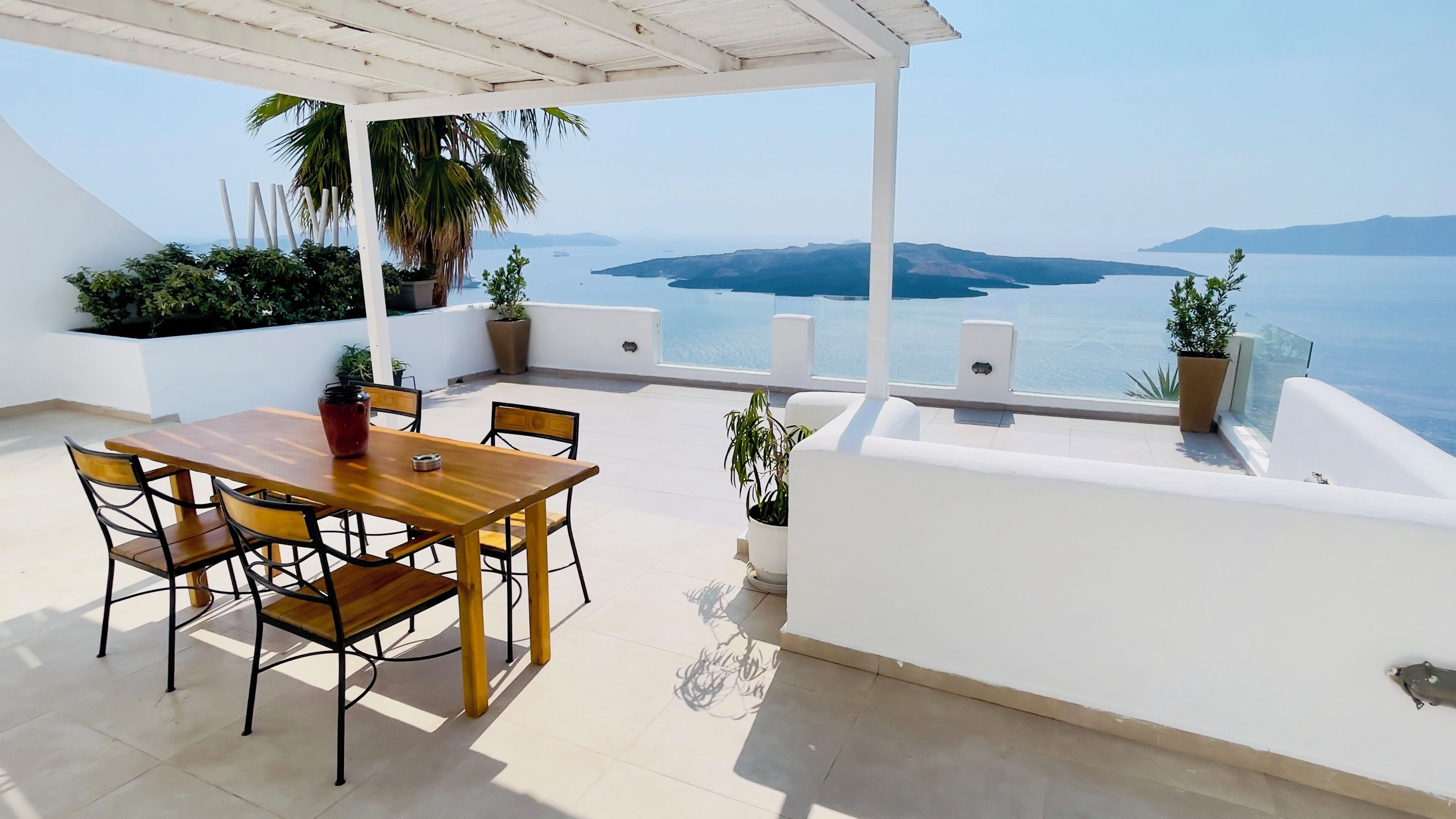 A terrace with a wooden table and chairs, potted plants, and a view of the sea.