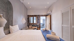 A bedroom with a large bed, a stone wall, a window, and a view of the city. - Loucas On The Cliff (Santorini)