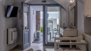 A hotel room with a TV, air conditioning unit, a blue towel, a white bed, a bathroom with a mirror, and a view of the outside balcony. - Loucas On The Cliff (Santorini)
