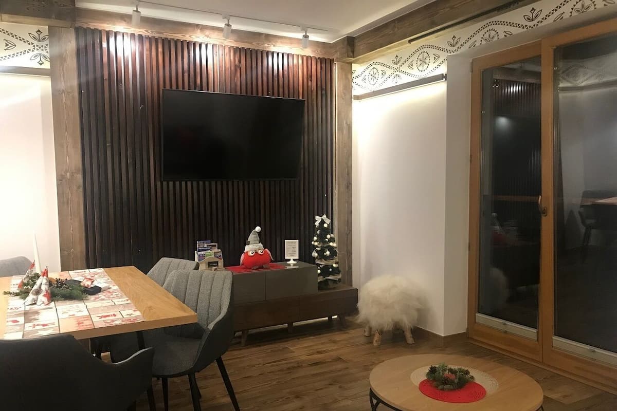 Uma sala de estar aconchegante com parede de madeira, uma TV de tela plana, uma mesa de jantar com cadeiras, uma pequena mesa redonda com um objeto decorativo e um cachorro branco e fofo.