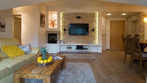 Een woonkamer met een stenen open haard, een flatscreen televisie aan de muur, een houten salontafel met fruit, een bank met kussens, en een eethoek met stoelen.