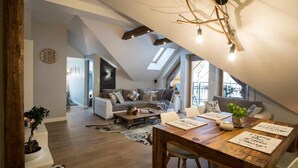 Een ruime loft met hoge plafonds, zichtbare houten balken, een eethoek met een houten tafel en een woonkamer met een bank en een open haard.