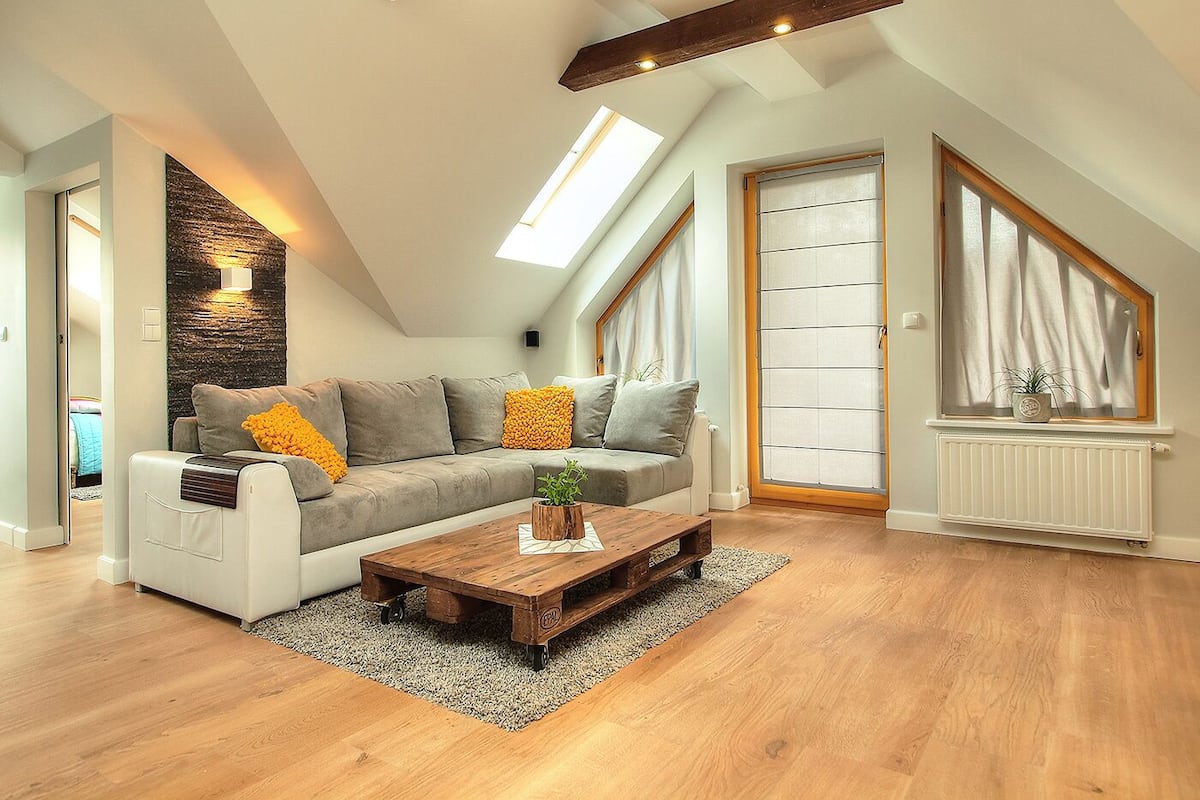 Uma sala de estar estilo loft com teto alto, vigas de madeira aparentes, uma janela grande, um sofá modular cinza e uma mesa de centro de madeira.