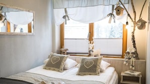 Een gezellige slaapkamer met een bed, kussens, een nachtkastje, een raam met gordijnen en een klein plankje met decoratieve objecten.