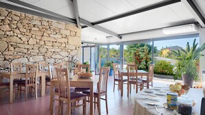 Comedor con mesas y sillas de madera, pared de piedra y ventanas amplias con vistas a zonas verdes y edificios.
