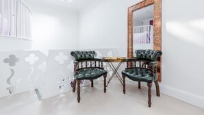 Ein Zimmer mit heller Einrichtung, einem Holztisch, zwei grünen Ledersesseln und einem großen Spiegel an der Wand.