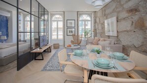 A modern living room with a stone wall, a round dining table set with plates and glasses, and a television on a stand. - Liiiving in Porto | Luxury Beachfront Apartment IV (Porto)