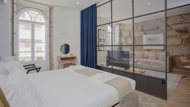 A modern hotel room with a large bed, a desk, a mirror, and a view of a balcony and living area through a glass partition. - Liiiving in Porto | Luxury Beachfront Apartment IV (Porto)