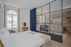 A modern hotel room with a large bed, a desk, a mirror, and a view of a balcony and living area through a glass partition.