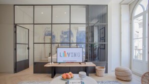 A modern living room with a glass partition, a mounted sign reading "LIVING," a round coffee table with books and a fruit bowl, and a view through the glass. - Liiiving in Porto | Luxury Beachfront Apartment IV (Porto)