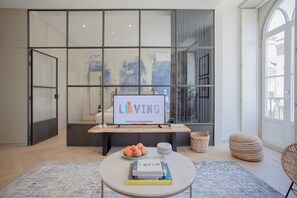 A modern living room with a glass partition, a mounted sign reading "LIVING," a round coffee table with books and a fruit bowl, and a view through the glass.