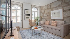 A living room with a stone wall, a sofa, a coffee table with fruit, and a wicker chair. - Liiiving in Porto | Luxury Beachfront Apartment IV (Porto)