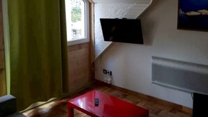 Une pièce avec un plafond en pente, un téléviseur fixé au mur, une table rouge et une fenêtre ornée d’un rideau vert.