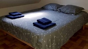 Un lit simple avec une couvre-lit grise à motifs et deux oreillers bleus, une lucarne au-dessus et un plancher en bois.