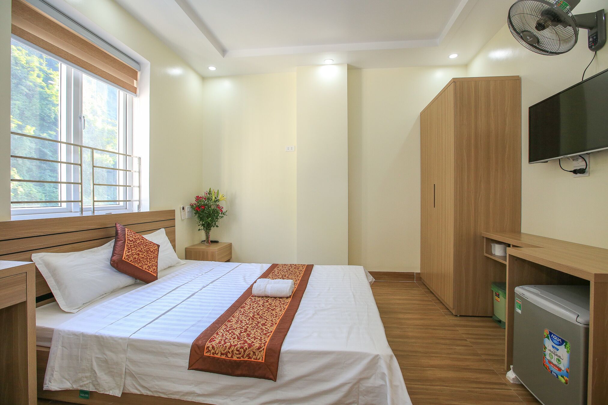 Una habitación de hotel con una cama, una mesita de noche, un televisor, un ventilador y un refrigerador pequeño.