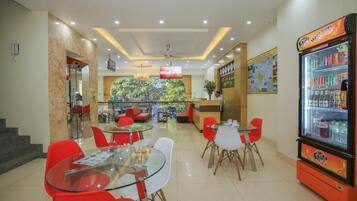 A hotel lobby with glass tables, red chairs, a vending machine, and a view of greenery through a glass wall.