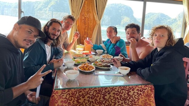 A group of people dining at a table with various dishes, a fan above, and a view of the outdoors. - Cat Ba Green Hotel (Hai Phong)
