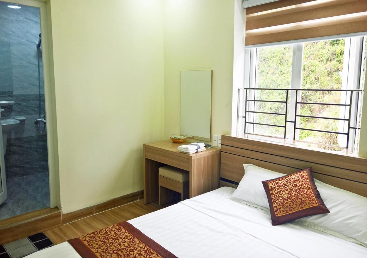 A hotel room with a bed, a bedside table, a mirror, a window with a view of greenery, and a shower area.