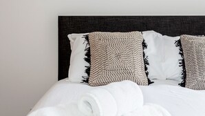 A neatly made bed with white linens and rolled towels, a framed artwork of a building on the wall, and a dark headboard.