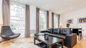 Ein modernes Wohnzimmer mit einer Ledersofa, einem grauen Sessel und einem Couchtisch. Große Fenster mit Vorhängen sorgen für natürliches Licht.