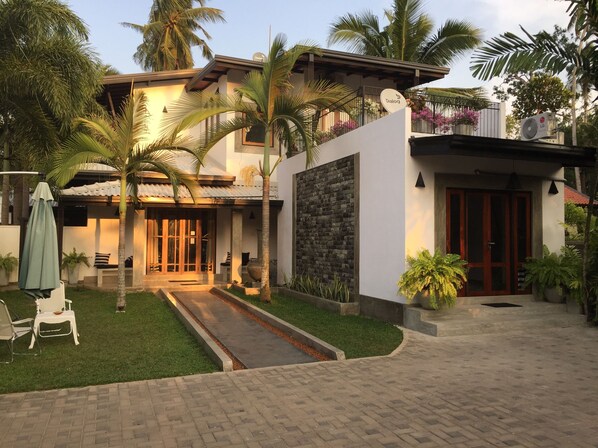 A modern house with a stone wall, a covered porch, and a well-maintained garden with palm trees and a pathway.