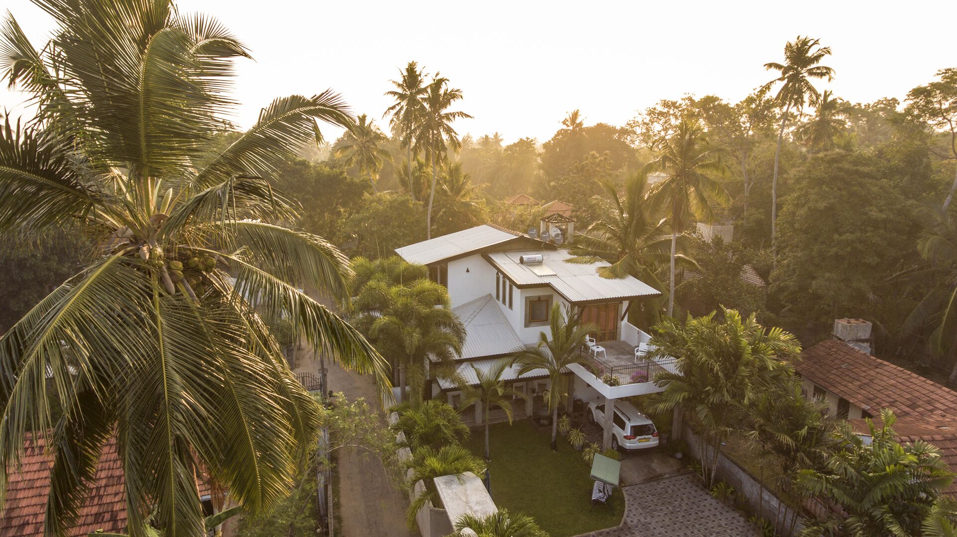 A two-story house with a flat roof, surrounded by lush greenery and palm trees.