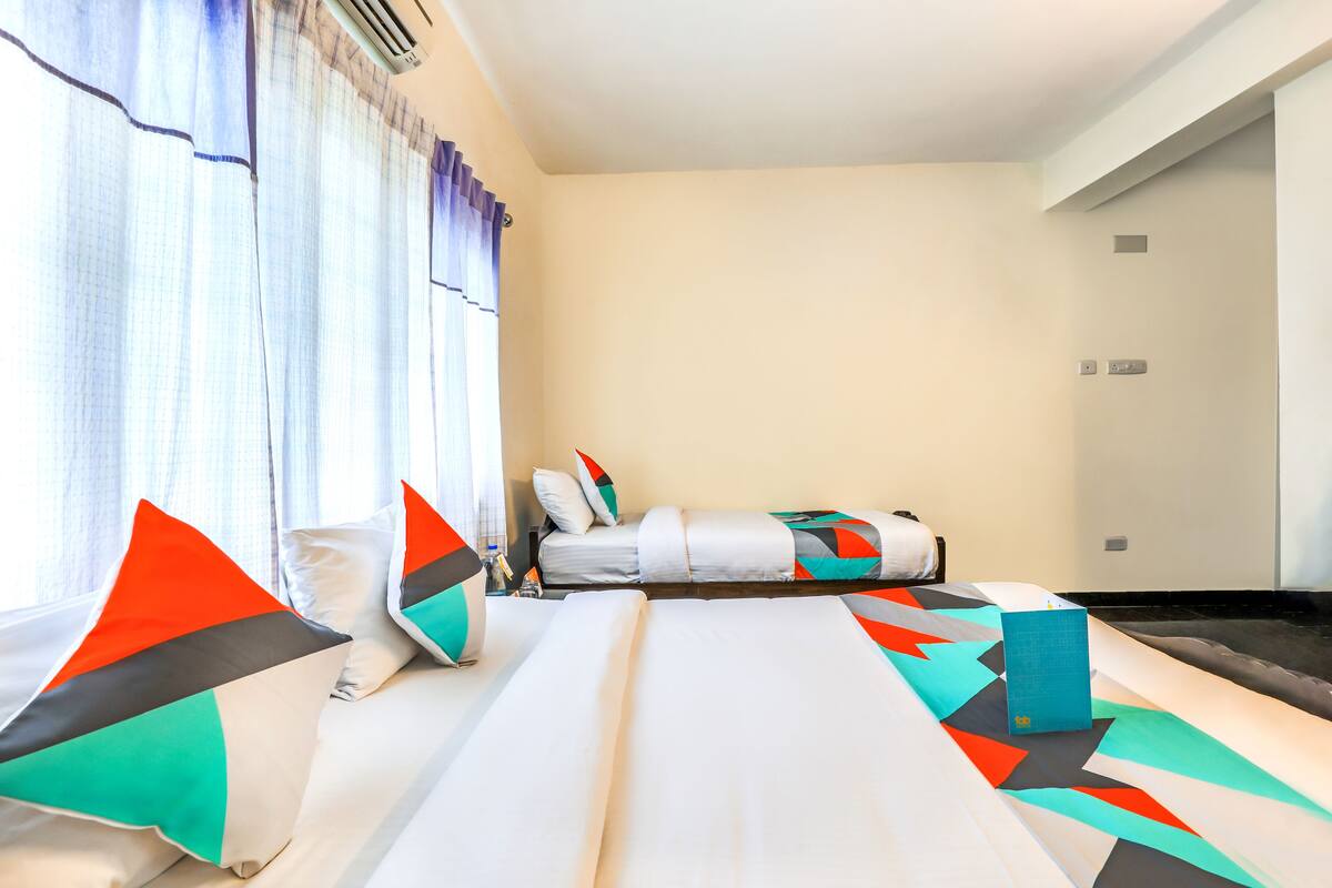 A hotel room with a large bed, a smaller bed, and a desk. There is a window with curtains, a wall-mounted air conditioner, and a small table with a bottle and glasses.