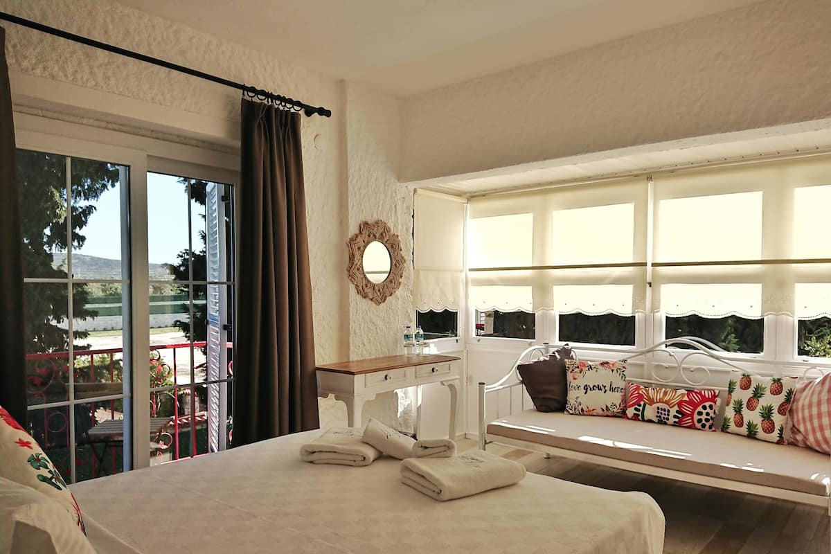 Habitación de hotel con cama, un banco con cojín floral, una mesita y una ventana con vistas al exterior.