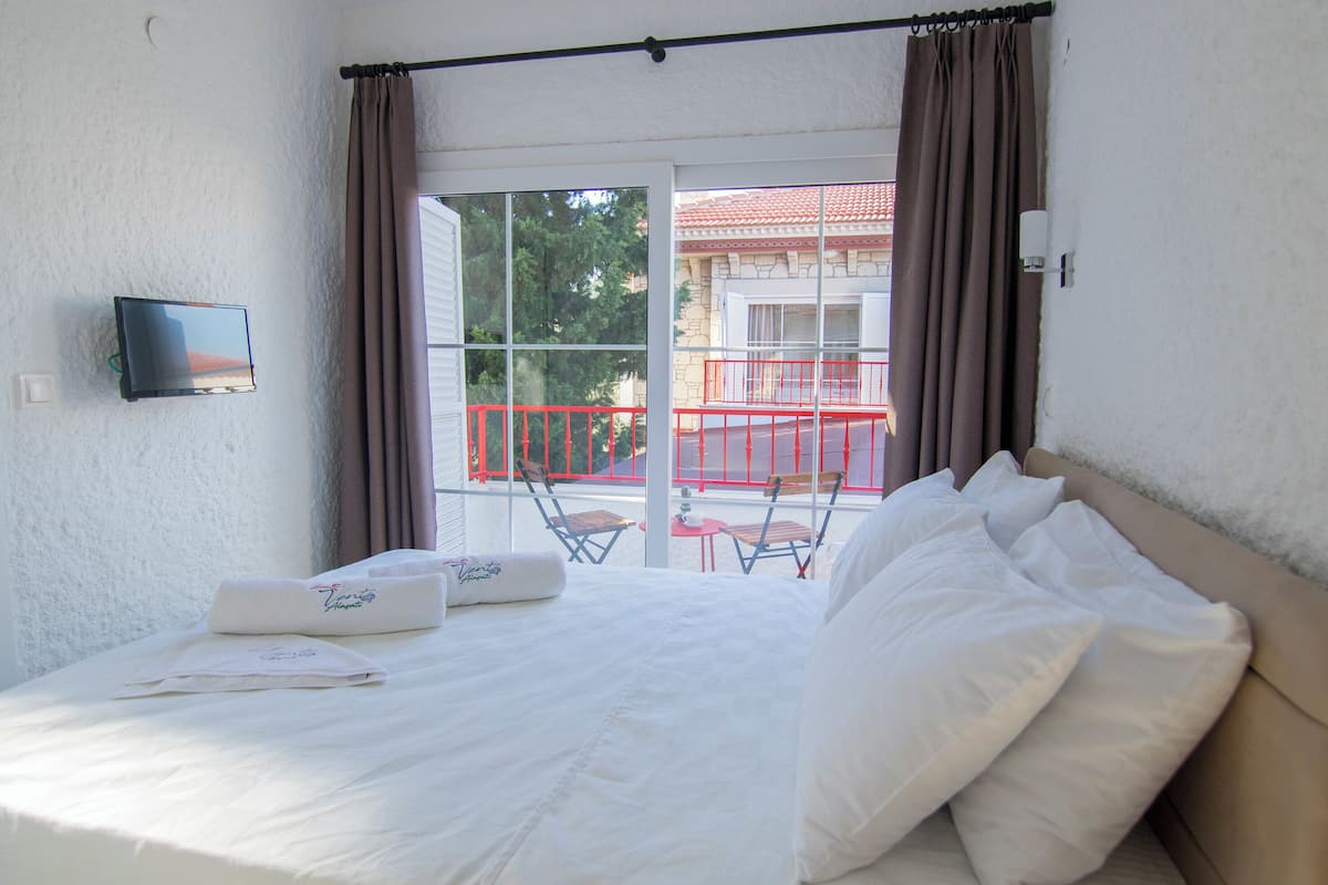 Habitación de hotel con cama, televisor montado en la pared y balcón con barandillas rojas.