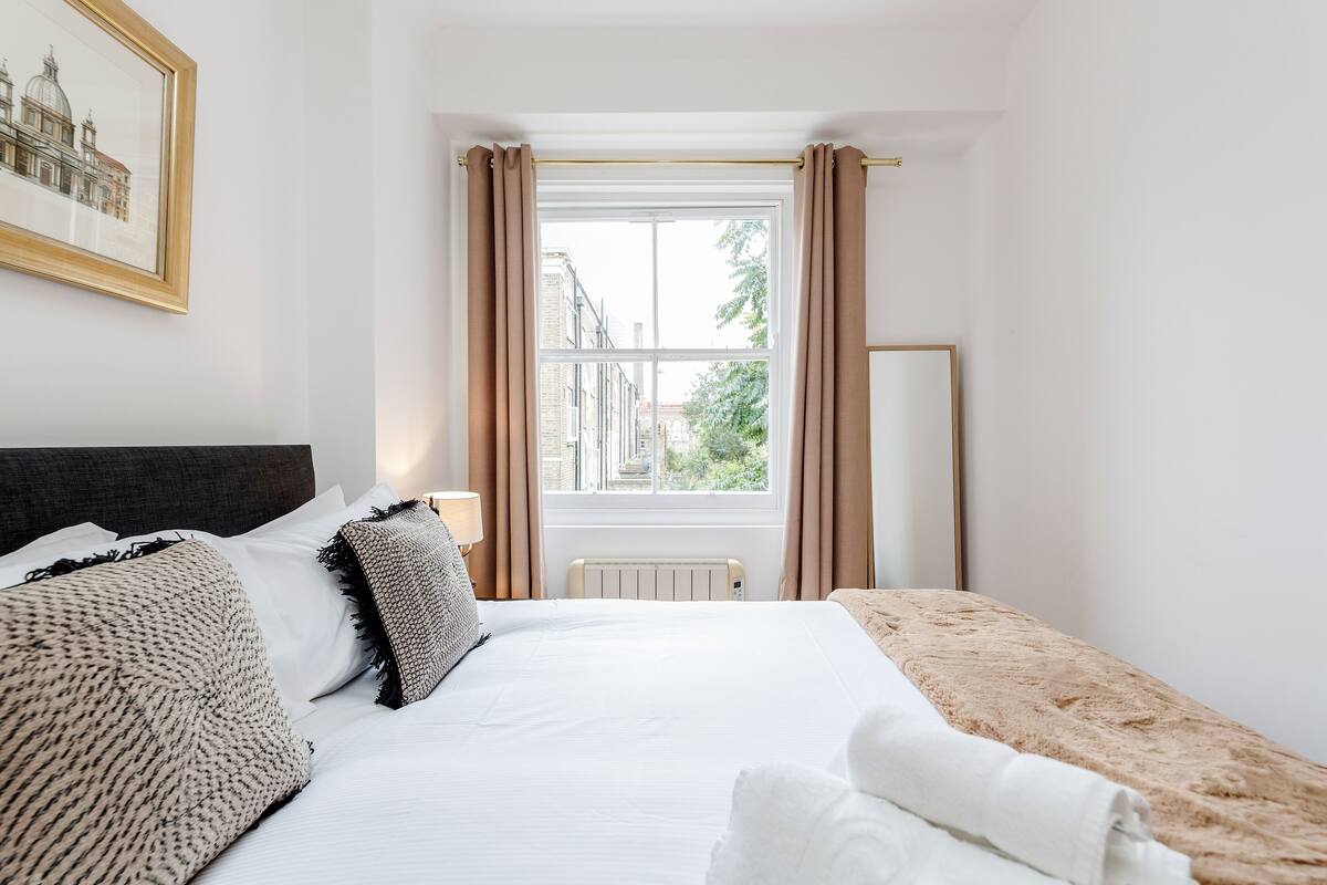 A hotel room with a large bed, a window with curtains, a radiator, and a framed picture on the wall.