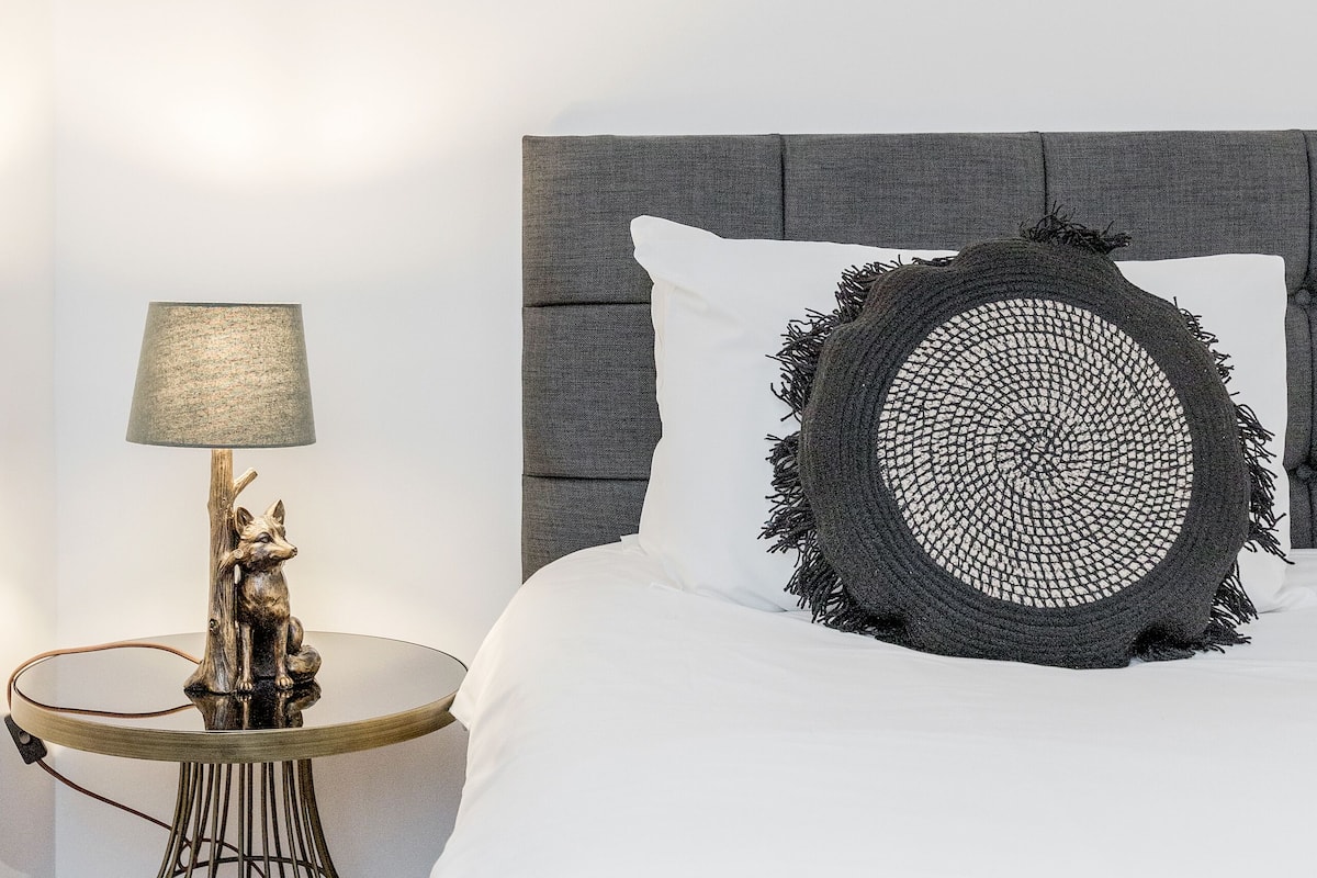 A neatly arranged bedroom with a bed, a round side table, a lamp, and a framed picture on the wall.