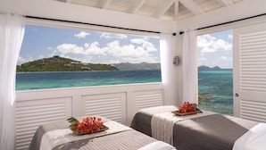 Une chambre avec deux lits, une vue sur la mer et une île au loin, et une fenêtre avec des volets blancs.