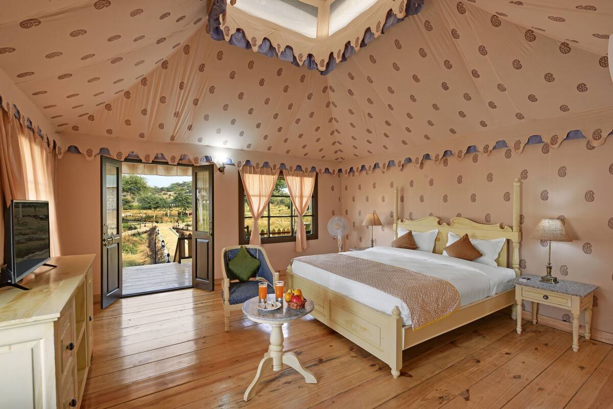 Een slaapkamer met een groot bed, een nachtkastje, een stoel, een klein tafeltje met fruit en drankjes, en een uitzicht door een open deur.