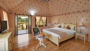A bedroom with a wooden floor, a bed with a headboard, a bedside table, a chair, a table with a tray of fruit and a glass of juice, and a view of the outdoors through an open door. - Jawai Leopard Safari Lodge (Sumerpur)