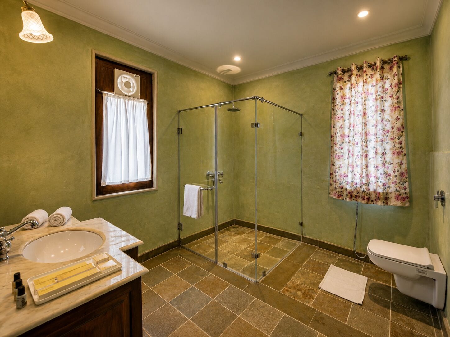 A bathroom with a glass-enclosed shower, a sink with toiletries, a toilet, and a window with curtains.