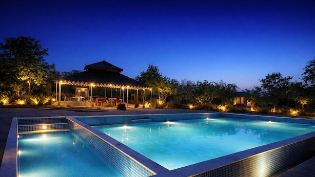 A swimming pool with tiled edges, surrounded by trees and a building with a covered patio. - Jawai Leopard Safari Lodge (Sumerpur)