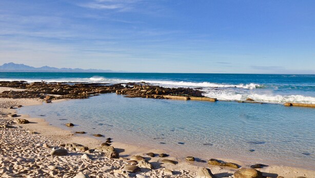 Una playa con agua azul cristalina, costa rocosa y montañas a lo lejos.