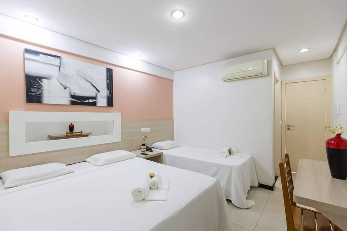 Habitación de hotel con dos camas, una mesita, una silla, un estante en la pared y un aire acondicionado.