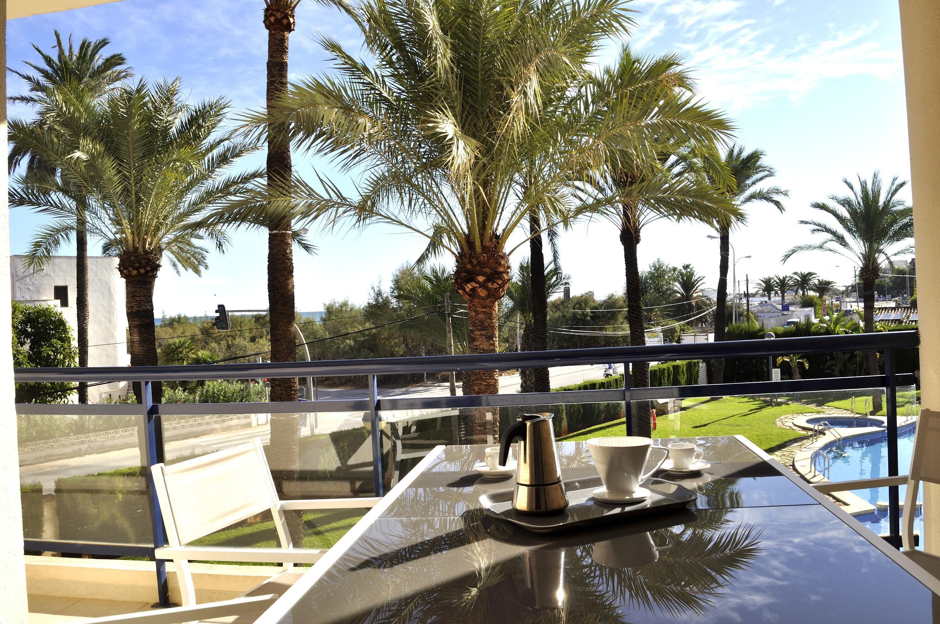 Un balcon avec une table, une cafetière et une tasse, donnant sur une piscine et des palmiers.