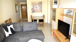 A living room with a grey sofa, a small white table, a TV on a wooden stand, and a dining area with a table and chairs. - Sueños De Denia, 2 bedrooms, by Bookindenia (Denia)