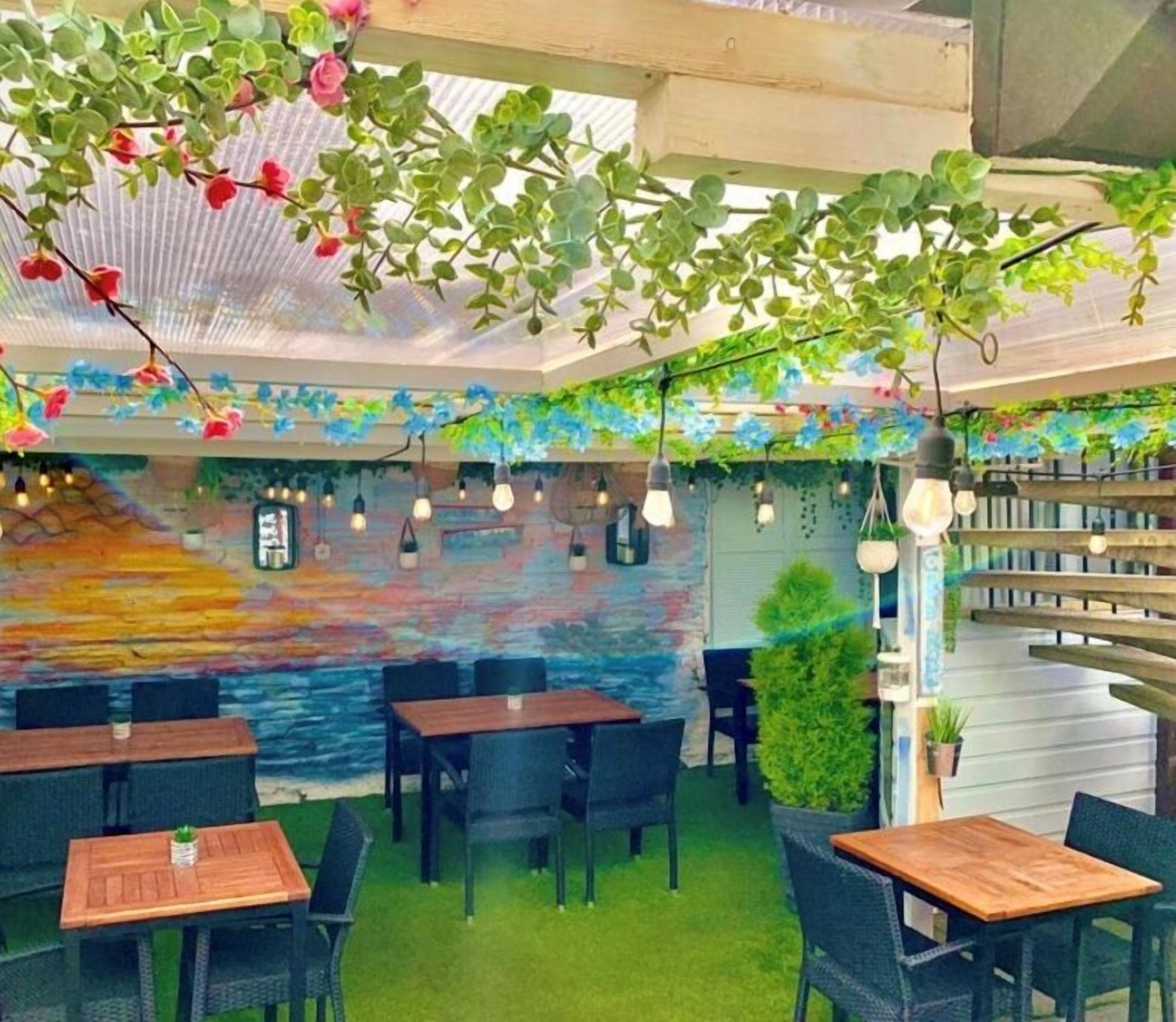 An indoor dining area with tables and chairs, a mural on the wall, hanging light bulbs, and green plants.