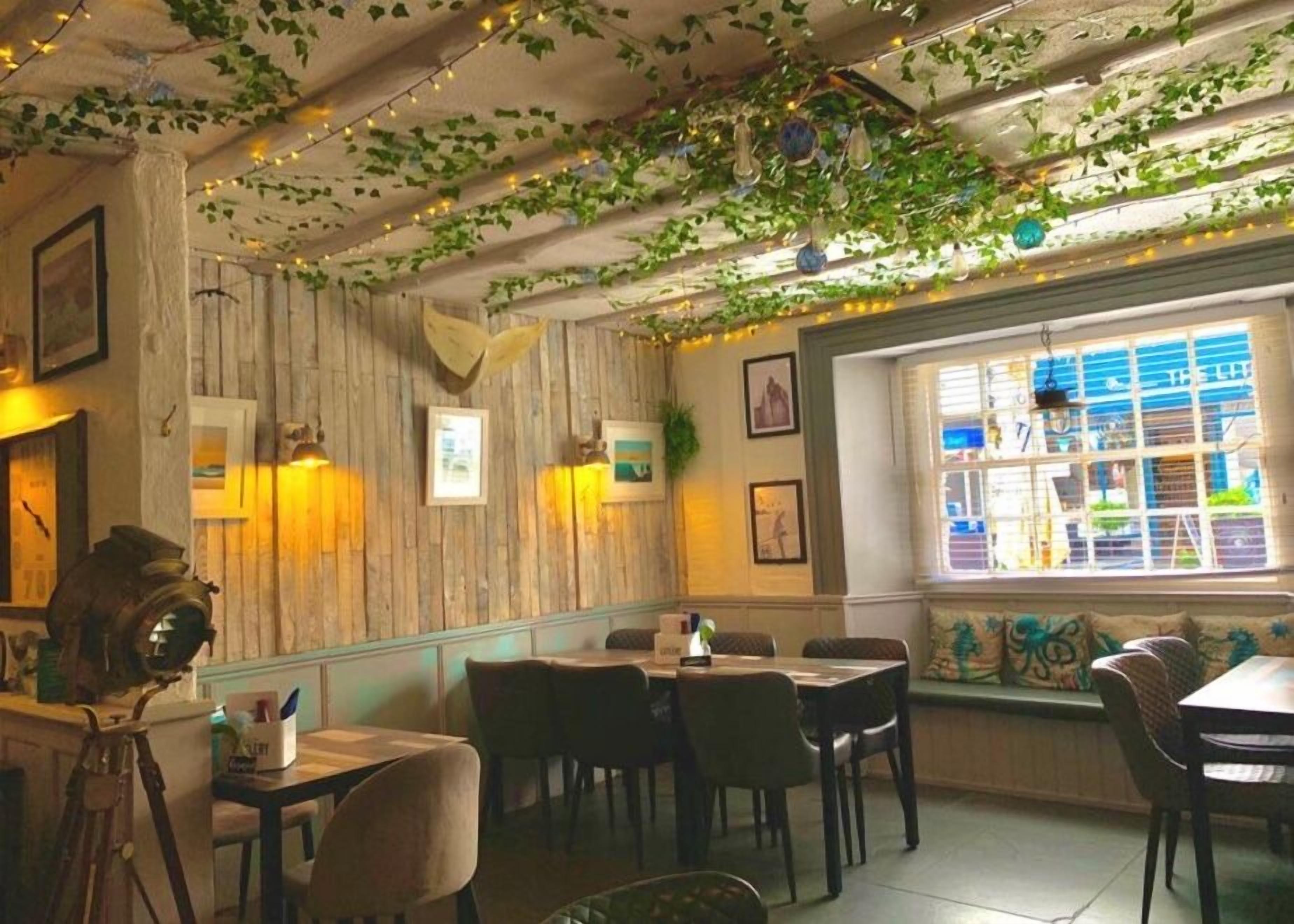 A restaurant with wooden walls, hanging plants, and string lights. There are tables and chairs for dining.