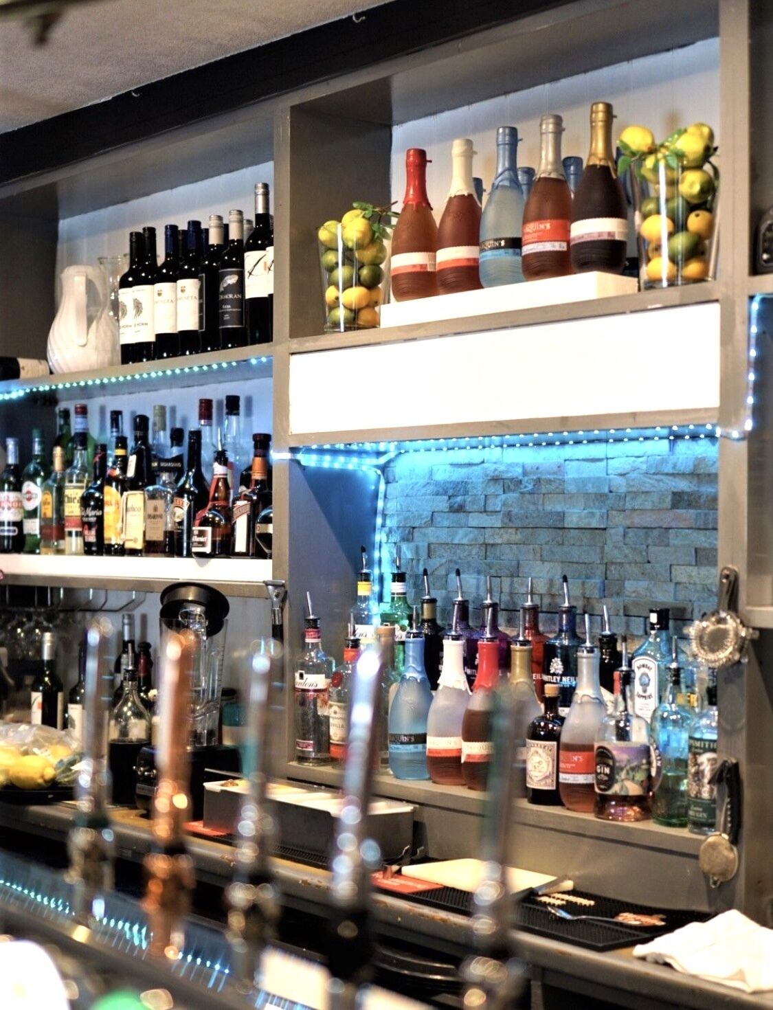 A bar with multiple shelves of liquor bottles, a brick wall, and a blue neon light.
