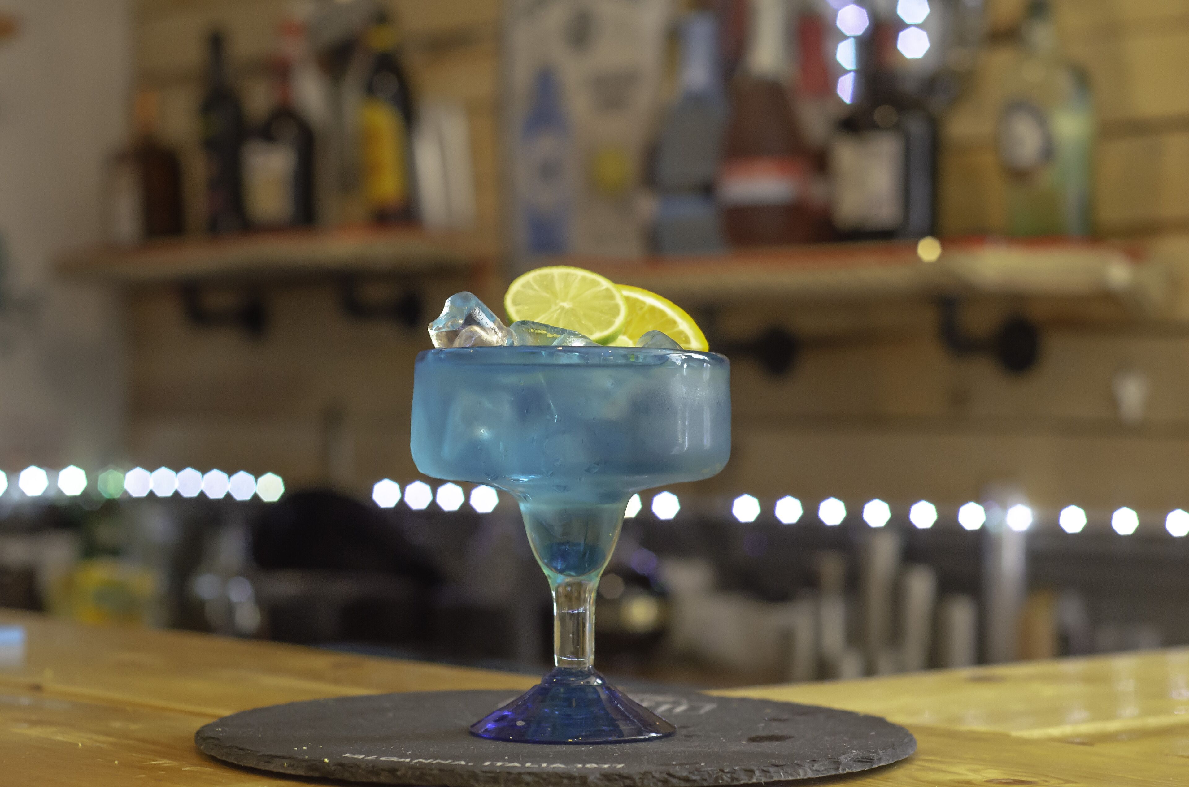 A cocktail glass with a blue drink, ice cubes, and a lime slice on a coaster.