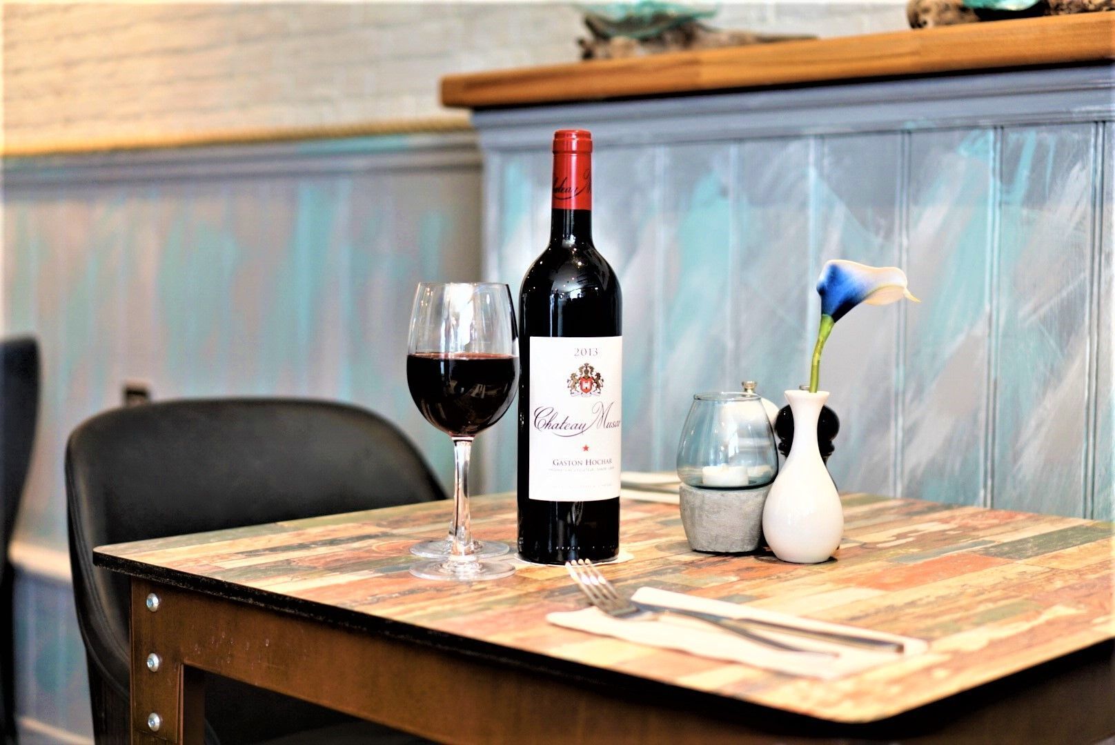 A table with a bottle of 2013 Chateau Mazar wine, a glass of red wine, a vase with a blue flower, and a candle holder.