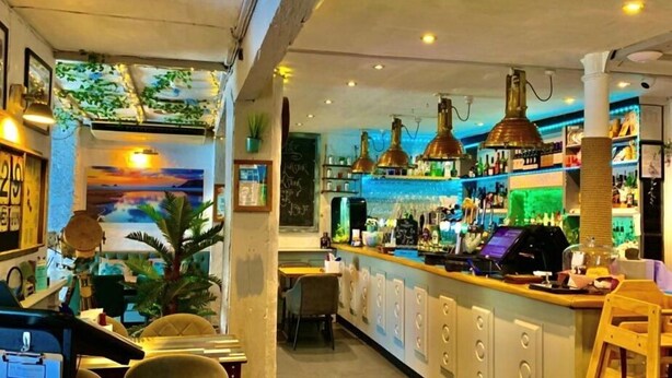 Eine Bar mit Theke, Barhocker und einer Auswahl an Flaschen. Pflanzen und eine Tafel mit der Speisekarte sind an der Wand angebracht.