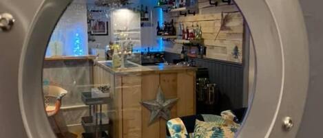 A circular window with a metallic frame, through which a bar area is visible, featuring a wooden counter, a star decoration, and various bottles.